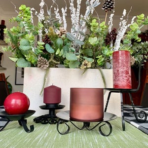 May include: A festive centerpiece with a white rectangular planter filled with greenery, pinecones, and berries. Several red candles of varying sizes are displayed on black metal candle holders, creating a warm and inviting holiday atmosphere.