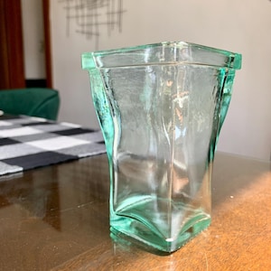 Puede incluir: Un jarrón de vidrio transparente de forma cuadrada con un ligero tinte verde. El jarrón tiene una superficie texturizada y una parte superior ligeramente ensanchada. Está sobre una mesa de madera, con un mantel a cuadros blanco y negro visible en el fondo.