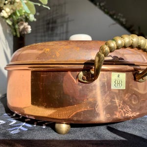 May include: A copper cooking pot with a brass handle and small brass feet. The pot has a white knob on the lid and a small label on the front. The pot is likely used for cooking and is a vintage or antique item.