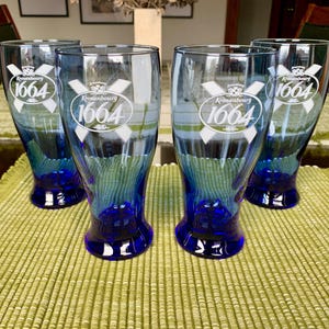 May include: Four blue glass beer mugs with a white logo that says "Kronenbourg 1664" etched on the front. The mugs have a blue base.