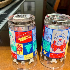 May include: Two clear glass drinking glasses with a Christmas theme. The glasses feature a patchwork design with red, green, blue, yellow, and white colors. One glass has a Mickey Mouse character wearing a Santa hat and the other glass has a snowman character. Both glasses have a snowflake design at the bottom.