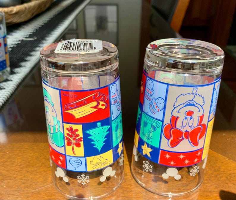 May include: Two clear glass drinking glasses with a Christmas theme. The glasses feature a patchwork design with red, green, blue, yellow, and white colors. One glass has a Mickey Mouse character wearing a Santa hat and the other glass has a snowman character. Both glasses have a snowflake design at the bottom.