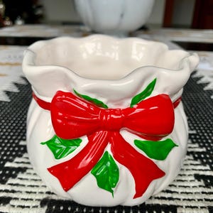 May include: White ceramic Christmas-themed planter shaped like a sack with a red bow and green holly leaves.