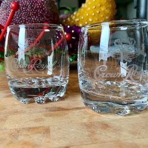 May include: Two clear, lowball glasses with the "Crown Royal" logo etched on the front. The glasses are filled with a clear liquid and sit on a wooden surface. The background includes colourful floral arrangements.