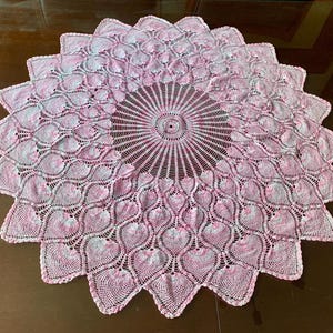May include: A round, handmade doily with a heart-shaped pattern in shades of pink and white. The doily has a central sunburst design and a scalloped edge. This decorative piece is perfect for adding a touch of elegance to any room.