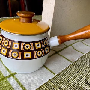 May include: Vintage white and orange enamel saucepan with a wooden handle and lid. The saucepan features a geometric pattern in brown, orange, and yellow. A classic kitchenware item.