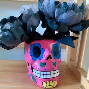May include: A pink ceramic skull planter with blue and green painted details. The skull has a painted smile and large blue eyes. The planter is decorated with black fabric flowers.