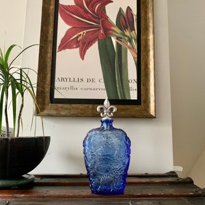 May include: A blue glass bottle with a decorative design of seashells and floral motifs. The bottle has a silver metal stopper with a fleur-de-lis design. The bottle is sitting on a wooden surface.