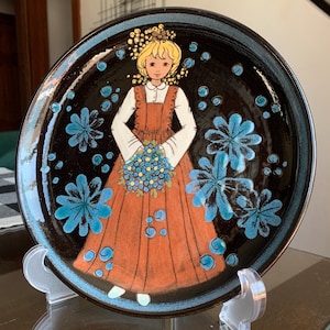 May include: A ceramic decorative plate with a black background and a blue rim. The plate features a stylized illustration of a girl in an orange dress holding a bouquet of blue flowers. Blue floral and dot designs are scattered across the plate.