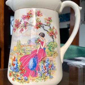 May include: A white ceramic mug with a black handle and the text "But First, Coffee" in a handwritten style. The mug is on a white surface. The mug is approximately 10 cm tall.
