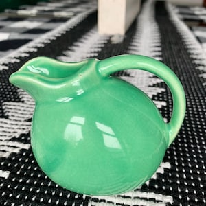 Peut inclure: Une petite cruche en céramique verte arrondie avec une poignée incurvée. La cruche a un bec verseur et une finition brillante. La cruche est posée sur une surface à motifs noirs et blancs.