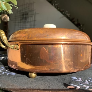 May include: A copper casserole dish with a lid and brass handles. The dish has a round shape and sits on three small brass feet. The lid has a white knob. The casserole dish is likely used for cooking and serving food.