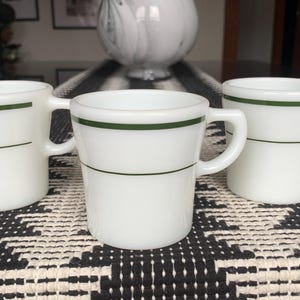May include: Three white ceramic mugs with a green stripe around the top. The mugs have a rounded shape and a handle on the right side.
