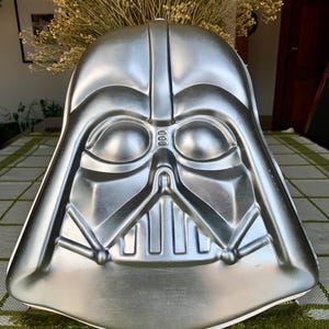 May include: A silver Darth Vader cake tin. The tin is shaped like the character's helmet, with detailed facial features. The tin is made of metal and is designed for baking cakes in the shape of the iconic Star Wars villain.
