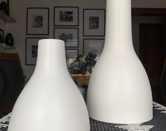 May include: Two white ceramic vases of different sizes. The larger vase is tall and slender, while the smaller vase is wider and shorter. Both vases have a smooth, rounded shape.