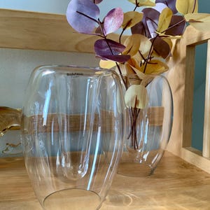 May include: Two clear glass vases with a textured, wavy design. The vases are filled with artificial eucalyptus leaves in shades of purple, yellow, and green. The vases are labeled "Boda Nova" in black text.