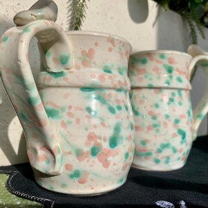 May include: Two ceramic mugs with a speckled design in pink and green on a white background. The mugs have a unique handle design and are suitable for beverages. The mugs are handmade and have a rustic, artistic appearance.