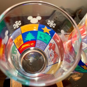 May include: Two clear glass cups with colorful Mickey Mouse themed designs. The designs include snowflakes, stars, and a variety of Mickey Mouse characters.