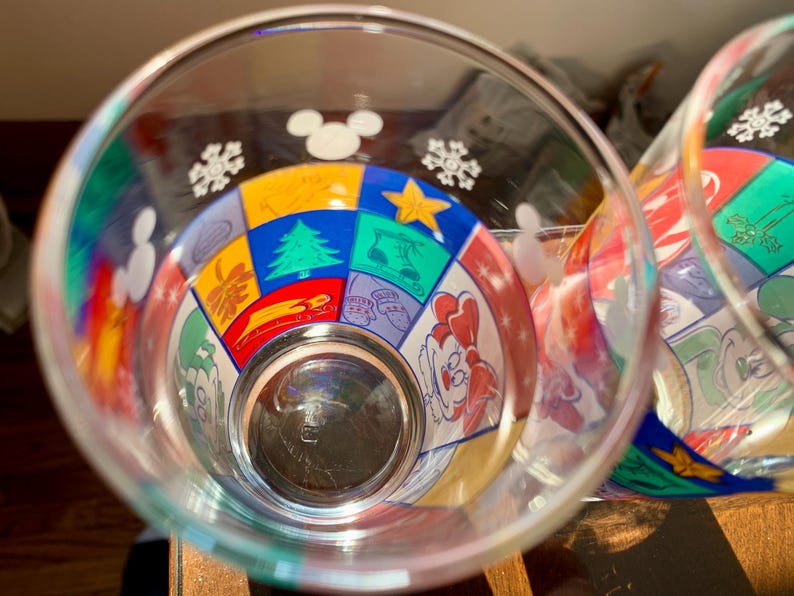 May include: Two clear glass cups with colorful Mickey Mouse themed designs. The designs include snowflakes, stars, and a variety of Mickey Mouse characters.