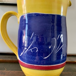 May include: A white ceramic mug with a black handle and the text "But First, Coffee" in a bold, black font. The mug is set against a blurred background, highlighting its clean design.