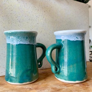 May include: Two handmade ceramic mugs featuring a vibrant teal green glaze with a contrasting white drip pattern around the upper rim. Each mug has a sturdy, curved handle and a smooth, glossy finish, showcasing a distinct, organic shape.
