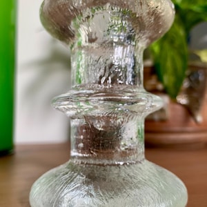 May include: A clear glass candlestick holder with a textured surface. The candlestick holder has a round base, a cylindrical stem, and a flared top. The glass has a slightly frosted appearance, and the overall design is simple and elegant.