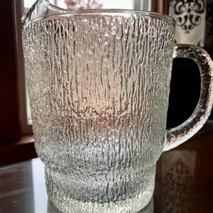 May include: A clear glass pitcher with a textured, bark-like surface. The pitcher has a handle and a spout for pouring. The design is reminiscent of tree bark. The pitcher is suitable for serving beverages.