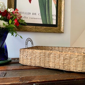 May include: A woven wicker basket with two metal handles. The basket is light brown and has a rectangular shape. The basket is sitting on a wooden surface.