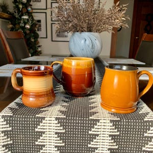 Puede incluir: Tres tazas de cerámica en tonos marrones y naranjas sobre un camino de mesa estampado. Las tazas tienen asas curvas y gradientes de color variables. Un jarrón con plantas secas se encuentra en el fondo.