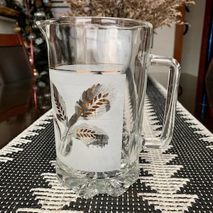 May include: A clear glass jug with a frosted white panel featuring a gold wheat design. The jug has a handle and a wide mouth.
