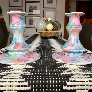 May include: Two ceramic candle holders with a colourful abstract design in shades of pink, blue, and green. The holders are on a black and white patterned table runner.