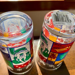 May include: Two clear glass drinking glasses with colorful cartoon characters and designs. The bottom of one glass has a sticker that reads "Hockin Glass Made in U.S.A." and a barcode.