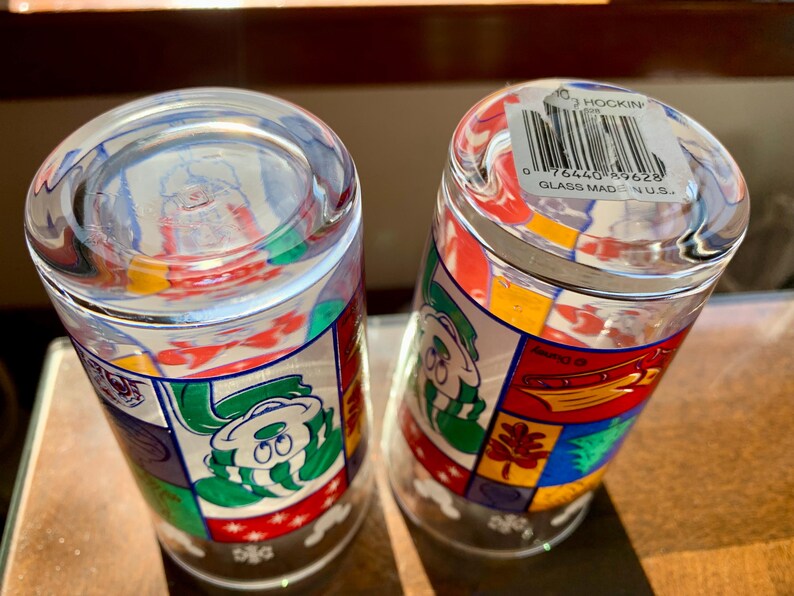 May include: Two clear glass drinking glasses with colorful cartoon characters and designs. The bottom of one glass has a sticker that reads "Hockin Glass Made in U.S.A." and a barcode.