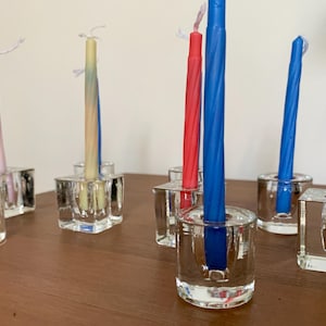 Può includere: Una collezione di candele colorate in portacandele di vetro trasparente. Le candele sono rosse, blu, rosa e gialle, con un design a spirale. I portacandele sono quadrati e cilindrici, disposti su una superficie di legno.