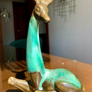 Peut inclure: Une figurine de cerf en céramique en position allongée avec un long cou dressé. La sculpture présente un vernis brillant avec un mélange de brun profond et de vert turquoise vif, créant une surface lisse et réfléchissante sur tout le corps.