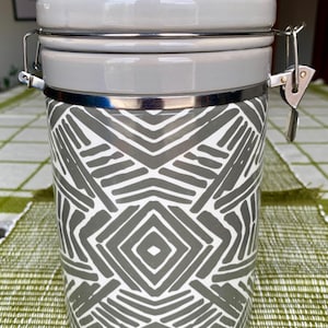 May include: A gray and white ceramic canister with a geometric pattern. The canister has a matching lid and a metal clasp closure. The design features a repeating pattern of gray lines and shapes on a white background. The canister is likely used for kitchen storage.