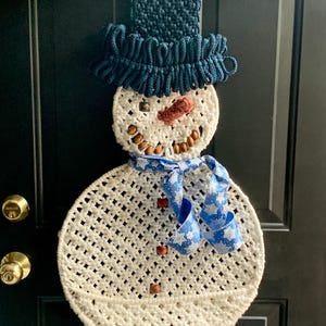 May include: A white crocheted snowman with a blue hat and a blue ribbon with white snowflakes. The snowman has a brown nose and brown eyes made of beads.