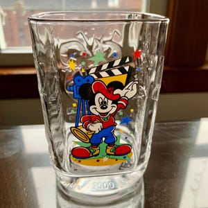 May include: A clear glass cup with a Mickey Mouse design. Mickey is wearing a red shirt, blue pants, and a red hat. He is holding a microphone and standing in front of a movie clapperboard. The cup has the year 2000 printed on the bottom.