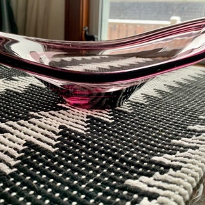 May include: A large, clear glass bowl with a deep purple hue. The bowl is shaped like a shallow dish with a wide, rounded base. It is sitting on a black and white patterned surface.