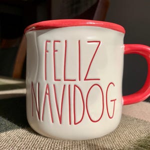May include: White ceramic mug with a red rim and handle. The mug features the text "FELIZ NAVIDOG" in red block letters. The mug is sitting on a textured surface.