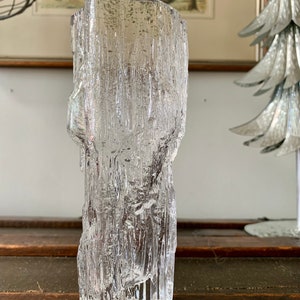 May include: A clear glass vase with a textured, bark-like exterior. The vase has a cylindrical shape with an uneven top edge. The glass appears to be thick and heavy, with a rough, organic texture. The vase is sitting on a dark wooden surface.