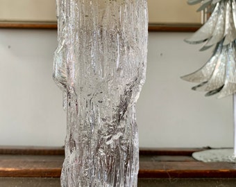 Peut inclure: Un vase en verre transparent avec un extérieur texturé en forme d'écorce. Le vase a une forme cylindrique avec un bord supérieur irrégulier. Le verre semble épais et lourd, avec une texture rugueuse et organique. Le vase est posé sur une surface en bois sombre.