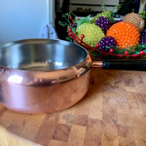 May include: A copper saucepan with a stainless steel interior and a black handle. The pan is on a wooden chopping board. A decorative bowl with colourful beaded spheres is in the background.