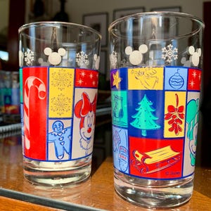 May include: Two clear glass drinking glasses with a Christmas theme. The glasses feature a patchwork design with various holiday icons, including Mickey Mouse, Minnie Mouse, a gingerbread man, a candy cane, a Christmas tree, and a stocking. The glasses are decorated with snowflakes and the Disney logo.