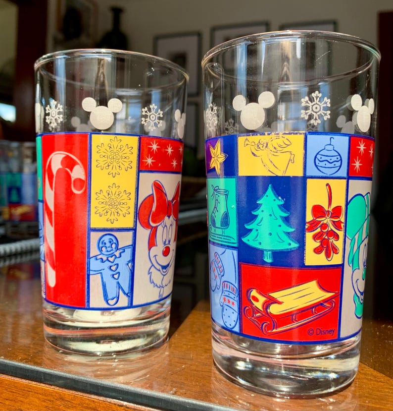May include: Two clear glass drinking glasses with a Christmas theme. The glasses feature a patchwork design with various holiday icons, including Mickey Mouse, Minnie Mouse, a gingerbread man, a candy cane, a Christmas tree, and a stocking. The glasses are decorated with snowflakes and the Disney logo.
