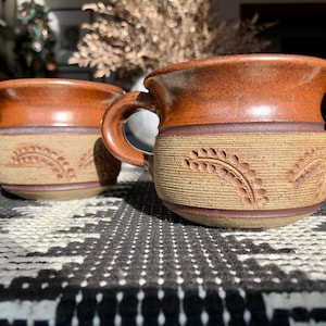May include: Two brown ceramic mugs with a textured design of wheat stalks. The mugs have a brown glaze and a light brown rim.