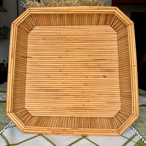 May include: A rectangular, light brown bamboo tray with a geometric design. The tray is made of thin, parallel bamboo rods, creating a textured surface. The edges are slightly angled, adding a unique shape. The tray is empty and sits on a patterned tablecloth.