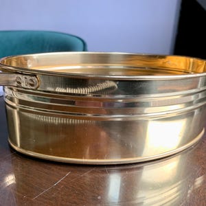 May include: An oval-shaped brass basin featuring a polished, reflective gold-toned finish. The container includes two flat, horizontal handles attached to the sides with visible rivets, and the body has a smooth, rounded rim and base.