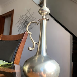 May include: A silver pewter jug with a bulbous body and a tall, pointed spout. The jug has a decorative handle and a shiny, reflective surface.