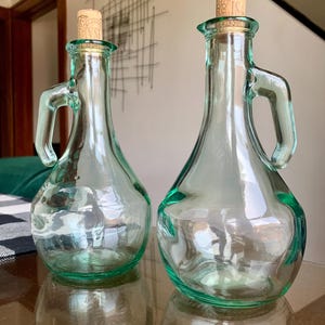 May include: Two clear, light green glass cruets with rounded bodies, narrow necks, and integrated side handles. Each vessel is topped with a cylindrical cork stopper featuring a stamped design. The glass has a slightly wavy, organic texture.